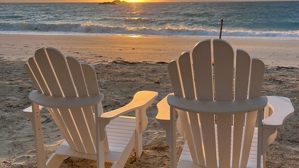 Deux chaises Adirondack blanches, placées face à l’océan, avec un coucher de soleil en arrière-plan.