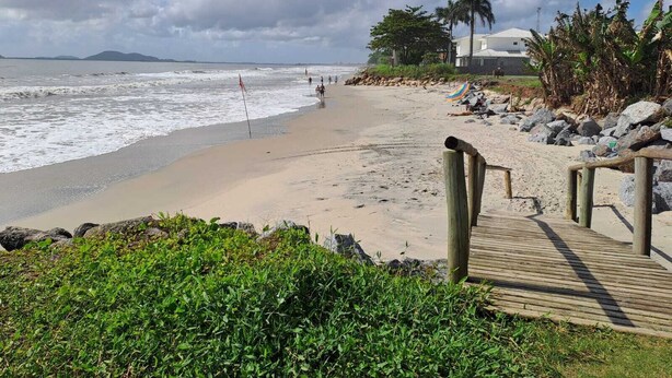 Una playa con un paseo marítimo de madera que conduce a la orilla, palmeras y un edificio al fondo.