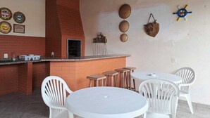 Un patio con una parrilla de ladrillo, sillas de plástico blanco y una mesa redonda.