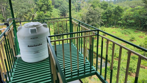 A balcony with a large white water tank labeled "Kelachandra" and a green metal railing overlooking a lush green landscape. - Forest home where elephants roam (Seethathode)