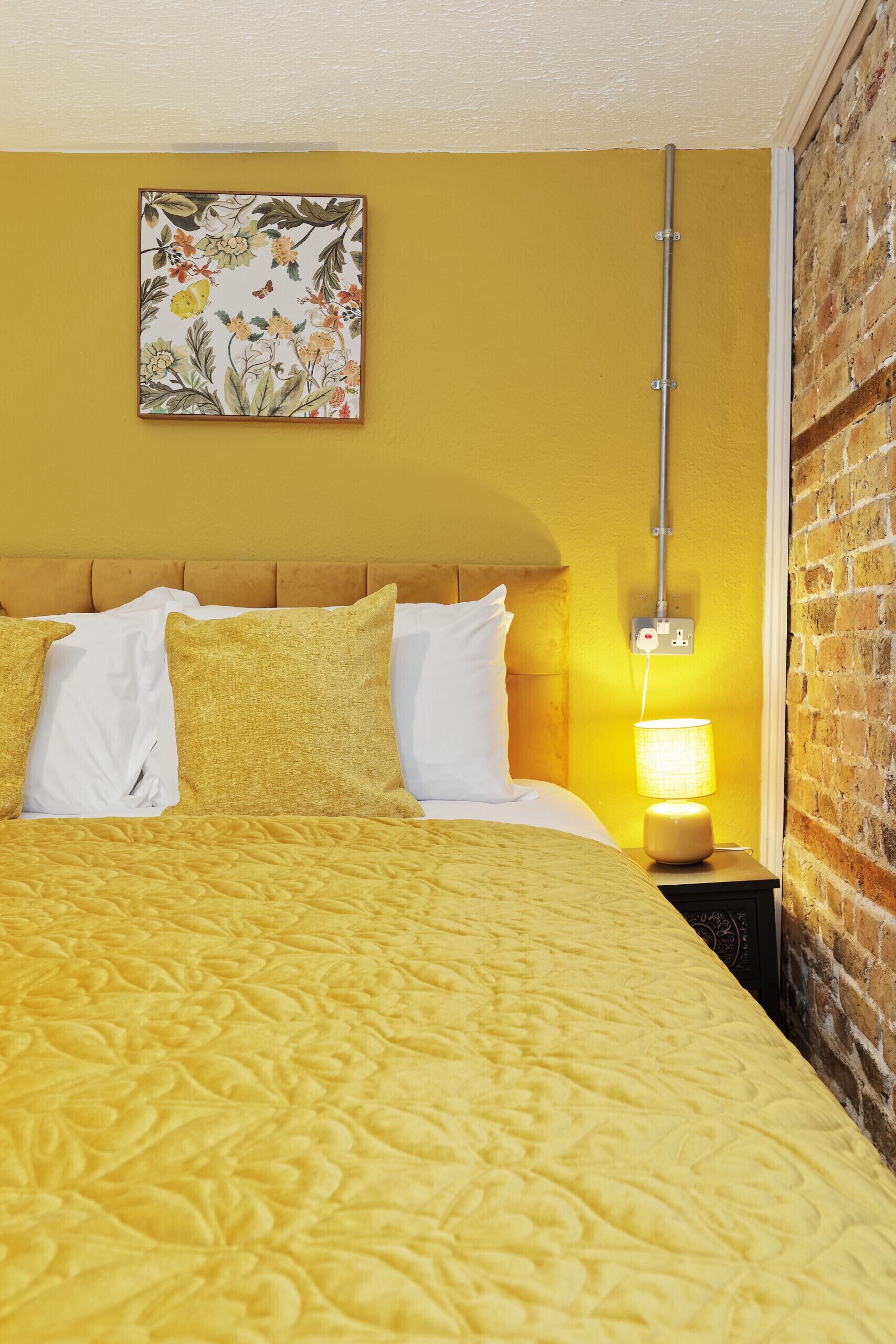 A room with a yellow bedspread, white pillows, a floral artwork on the wall, a nightstand with a lamp, and a brick wall.