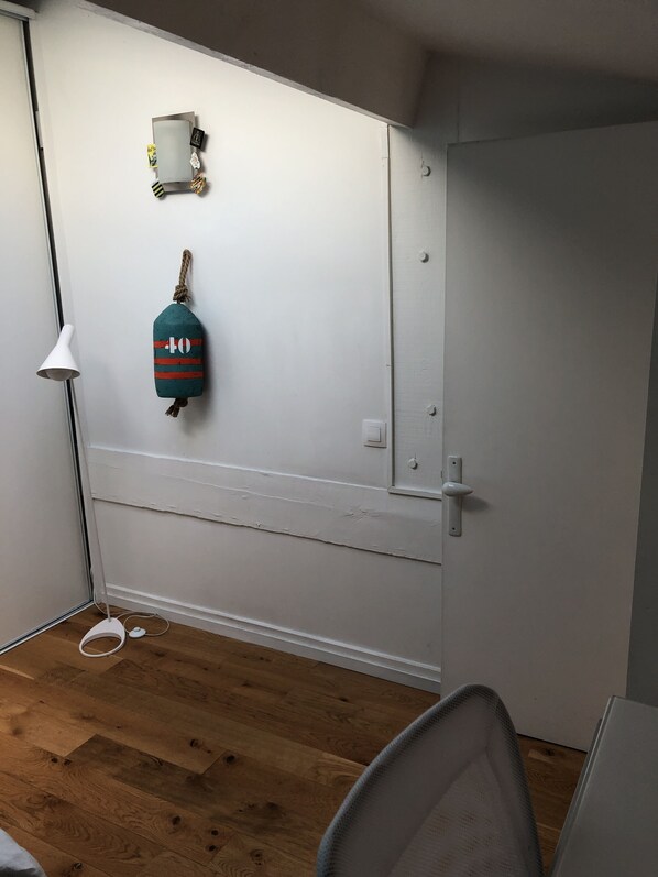 Una habitación con una puerta blanca, una pared blanca con un estante pequeño, un adorno colgante color turquesa y una lámpara blanca.