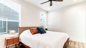 Un dormitorio con cama, mesita de noche, lámpara, ventilador de techo y persianas.