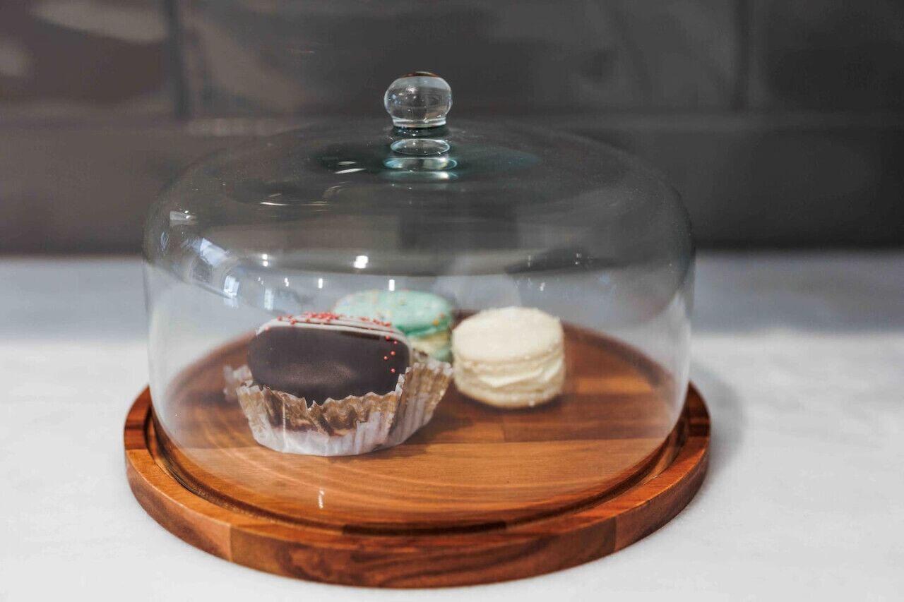 A dessert display with a glass dome over three macarons on a wooden board.