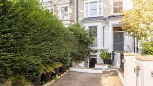 Een woonomgeving met een betegeld terras, diverse beplanting en een wit huis met een donkere voordeur.