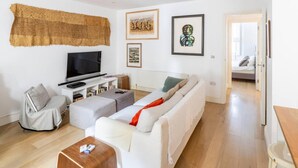 Een moderne woonkamer met een bank, een voetenbankje en een tv op een standaard. Er hangen kunstwerken in lijsten aan de muur en een deur leidt naar een andere kamer.