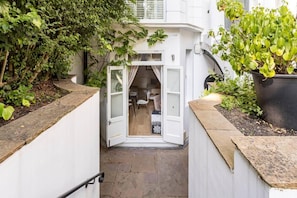 Un étroit sentier mène à une petite pièce fermée, dotée d’une porte, et entourée de verdure ainsi que d’une plante en pot.