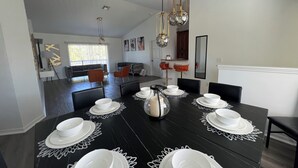 Ein Essbereich mit einem schwarzen Tisch, gedeckt für sechs Personen, weißen Tellern und einer Karaffe. Orangefarbene Stühle, eine Couch und eine Küche sind im Hintergrund zu sehen.