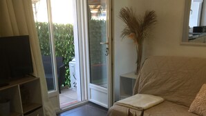 A living room with a sofa, a coffee table with decorative items, a television, and a sliding glass door leading to a patio. - Joli Studio rdc Avec Jardin Privé et Ombragé (LE CANNET DES MAURES)