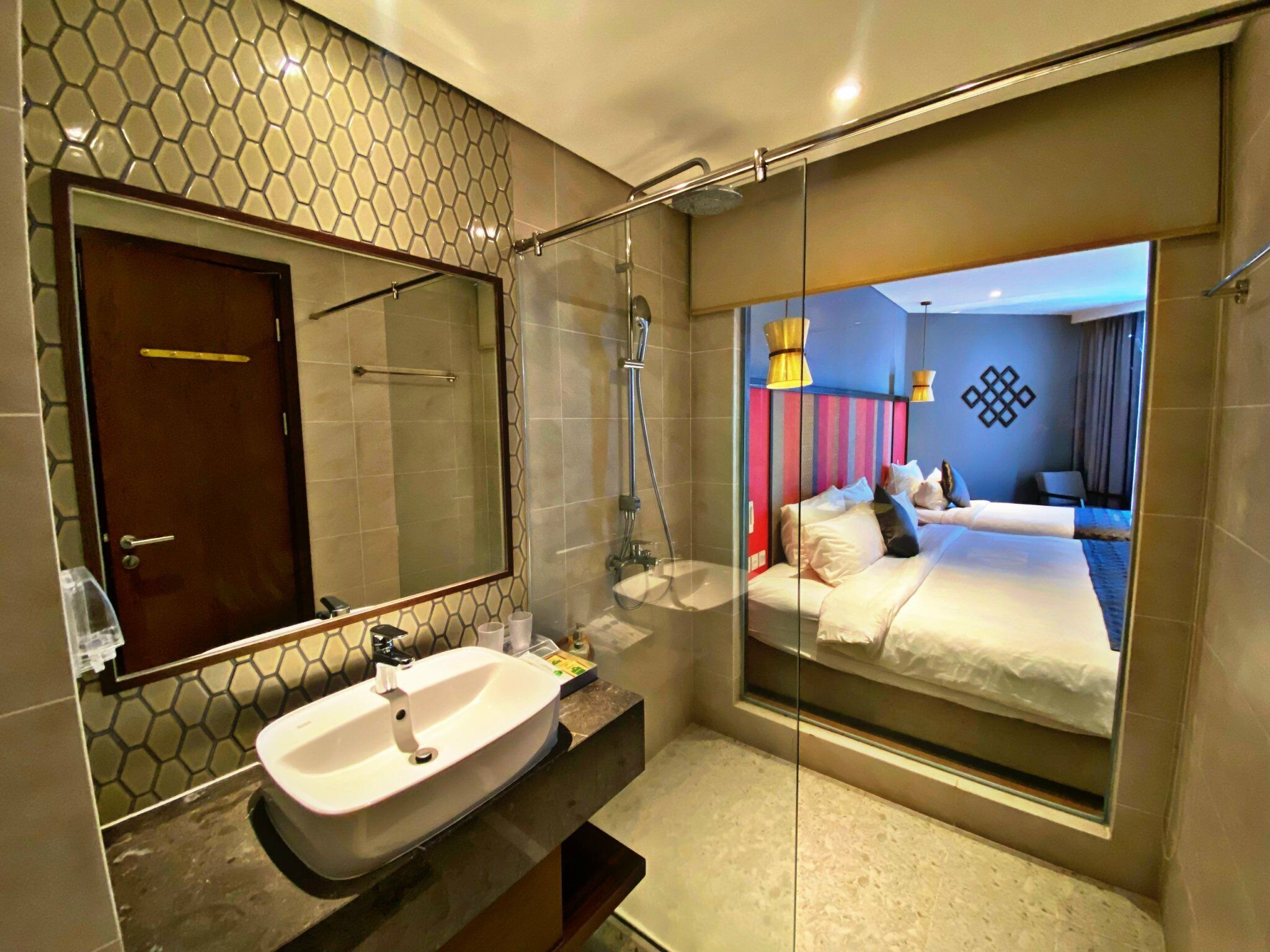A modern bathroom with a large mirror, a glass shower enclosure, and a visible bedroom with a bed and a nightstand.