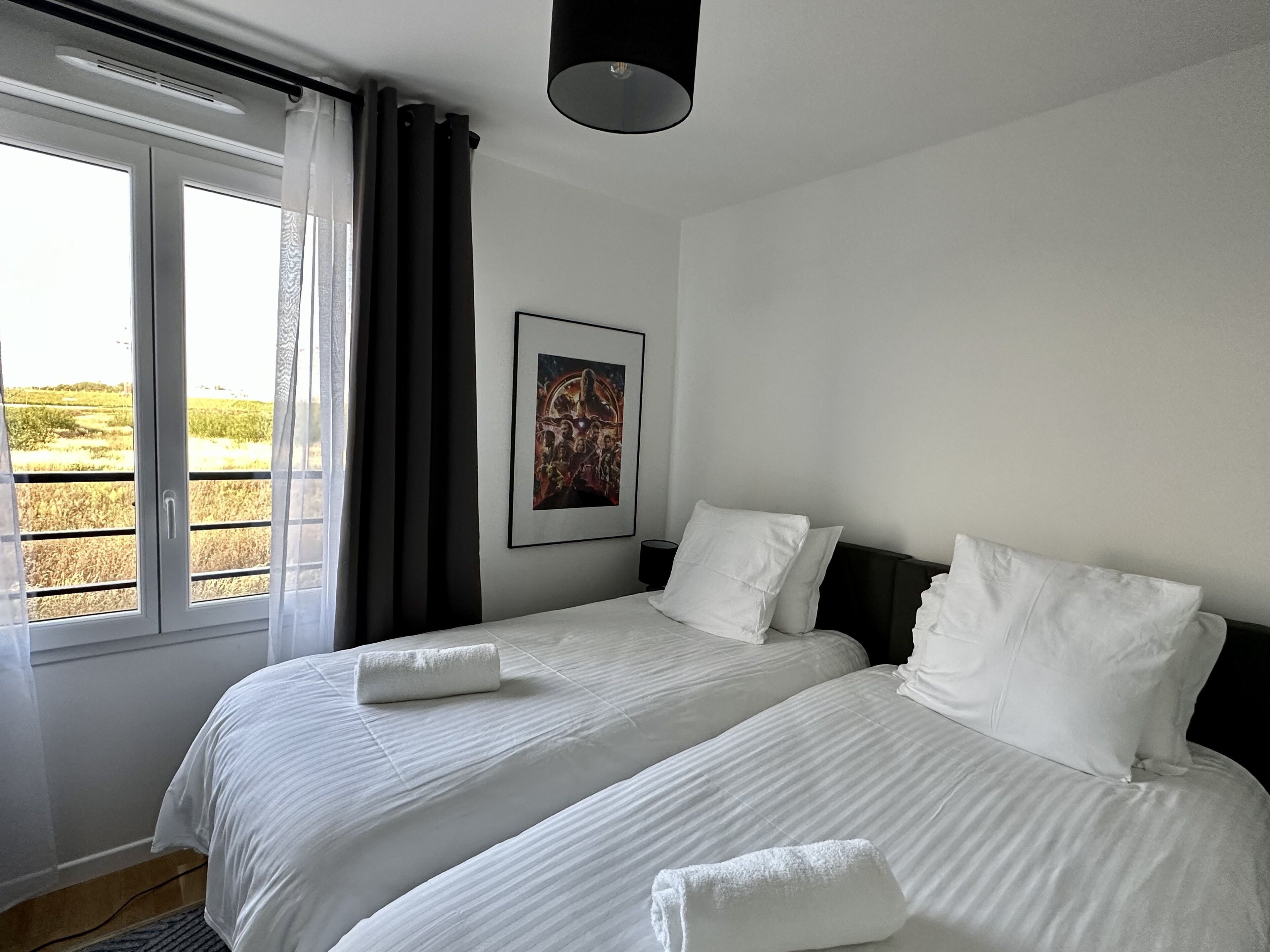 A hotel room with two beds, a large window with curtains, a framed picture on the wall, and a view of the countryside.