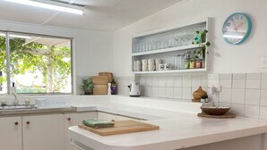Une cuisine moderne avec des meubles blancs, un îlot central, une fenêtre donnant sur un espace verdoyant et une horloge au mur.