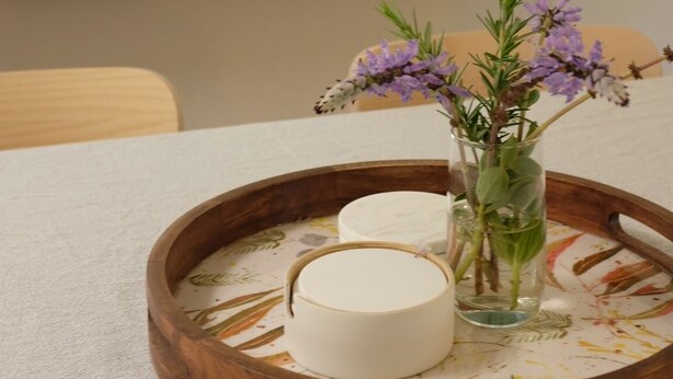 Un plateau en bois avec une bougie blanche et un vase en verre contenant des fleurs violettes, le tout posé sur une table.