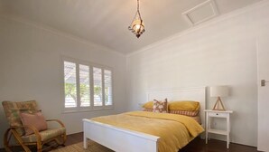 Une chambre à coucher avec un lit blanc, une chaise en osier, une table de chevet et une fenêtre avec des stores.