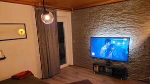 Ein Wohnzimmer mit Fernseher, einer Couch, einem kleinen runden Tisch, einer Fernbedienung und einer Wand mit Steinoptik.