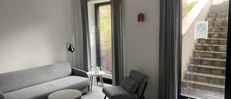 A modern living room with a grey sofa, a grey armchair, and a small round ottoman. There is a window with grey curtains, a small table with a plant, and a staircase visible outside.