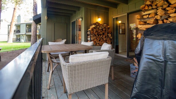 A covered outdoor seating area with wicker chairs and a table, a stack of firewood, and a view of a building in the background.