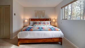 A bedroom with a wooden bed, two bedside lamps, a macrame wall hanging, and a window with a view of trees.