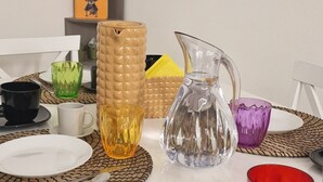 A dining area with a white table set for two, featuring a black cup, a clear glass pitcher, and various colorful glassware.