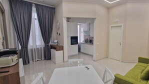 A modern living room with a dining table, a green sofa, and a kitchen area visible through an open door.