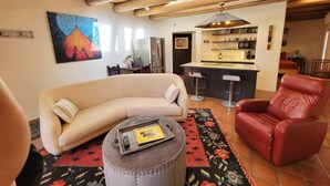 Een zithoek met een bank, poef en rode fauteuil. In de achtergrond is een keuken met een bargedeelte en een koelkast zichtbaar.