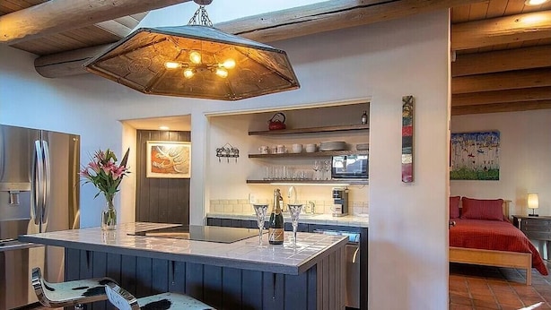 Een moderne keuken met een centraal eiland, barkrukken, een hanglamp en een rood bed in de aangrenzende kamer.
