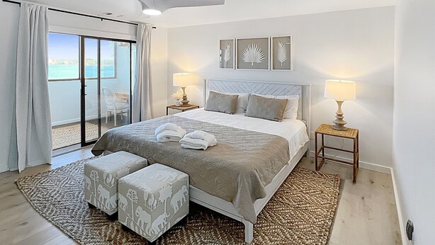 A bedroom with a bed, two bedside tables, a ceiling fan, and a view of the sea through a sliding door.