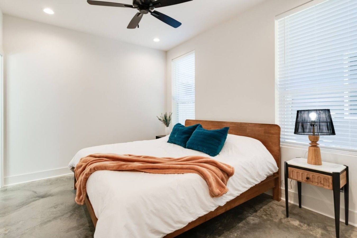 A bedroom with a bed, a ceiling fan, a nightstand with a lamp, and a window with blinds.