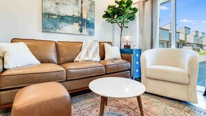 A living room with a brown leather sofa, a white armchair, and a small white table. There is a large abstract painting on the wall and a potted plant.