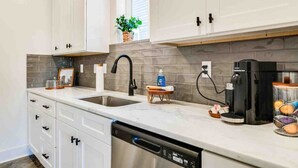 A modern kitchen with white cabinets, a gray backsplash, a dishwasher, a coffee maker, and a small plant on the countertop.