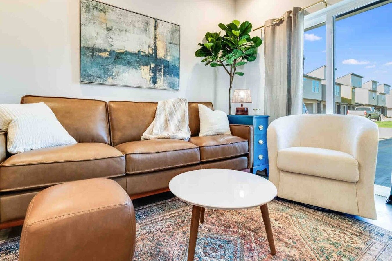 Un salon avec un canapé en cuir marron, une table basse blanche et un fauteuil beige. Un grand tableau abstrait orne le mur, et une plante en pot est posée à proximité.
