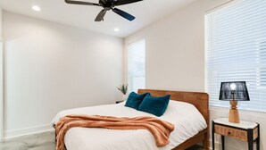 A bedroom with a bed, a ceiling fan, a nightstand with a lamp, and a window with blinds.