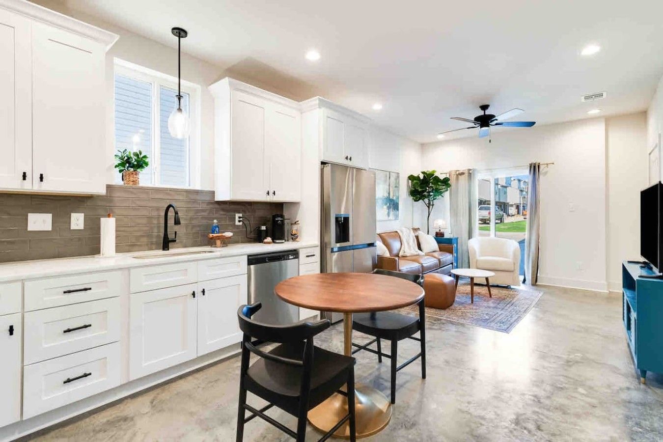 A modern kitchen with white cabinets, a dining area with a round table and chairs, and a living room with a sofa and a TV.