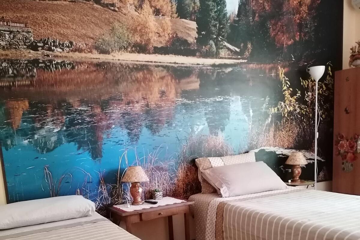 Une chambre avec deux lits, une table de chevet, une lampe et une fresque représentant un lac.