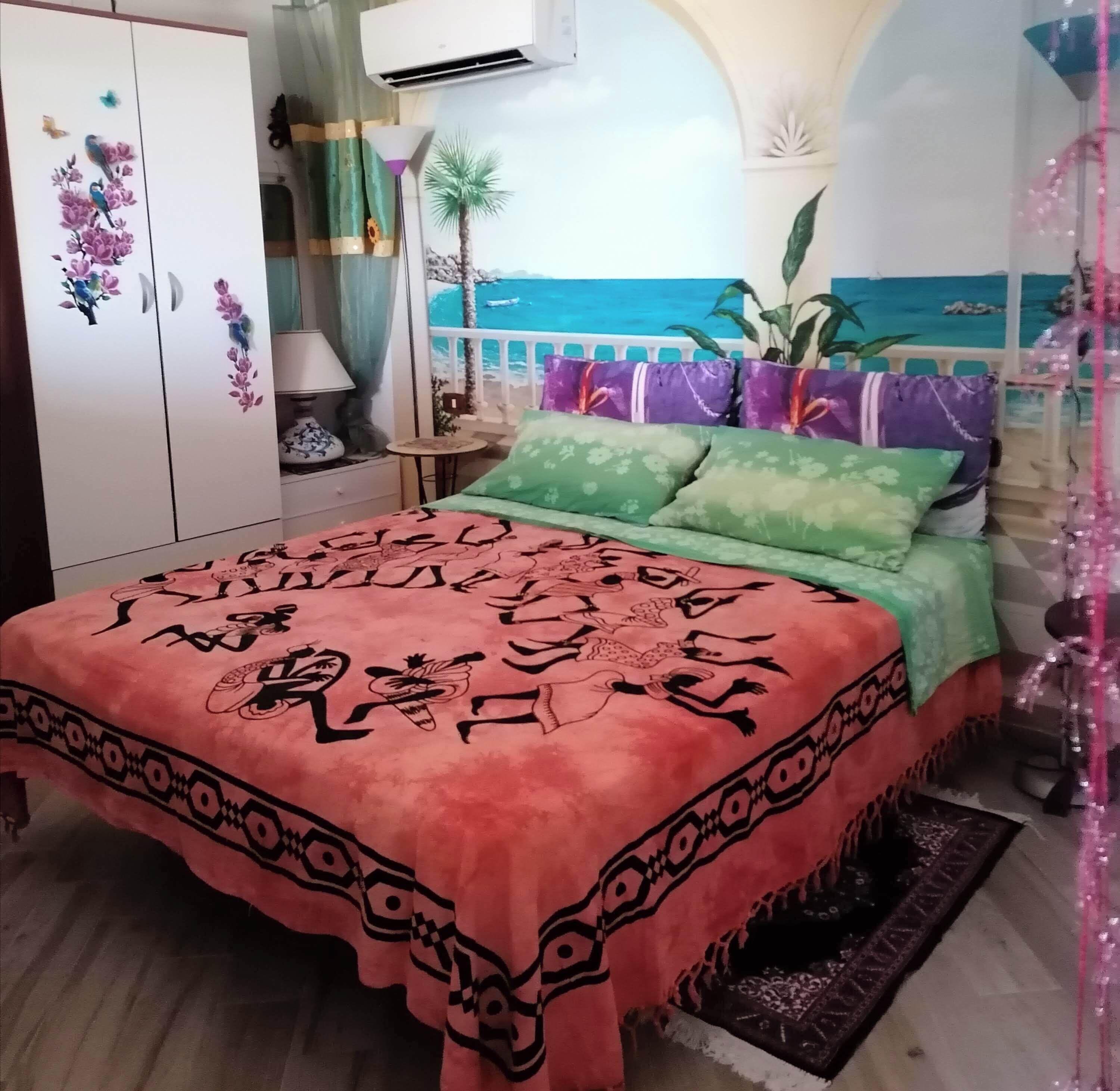 Een slaapkamer met een bed, een kledingkast, een nachtkastje, een lamp en uitzicht op zee door een raam.