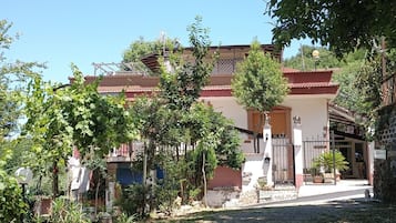 Un bâtiment avec un toit rouge, une entrée fermée par un portail et un petit jardin agrémenté d’arbres et d’arbustes.