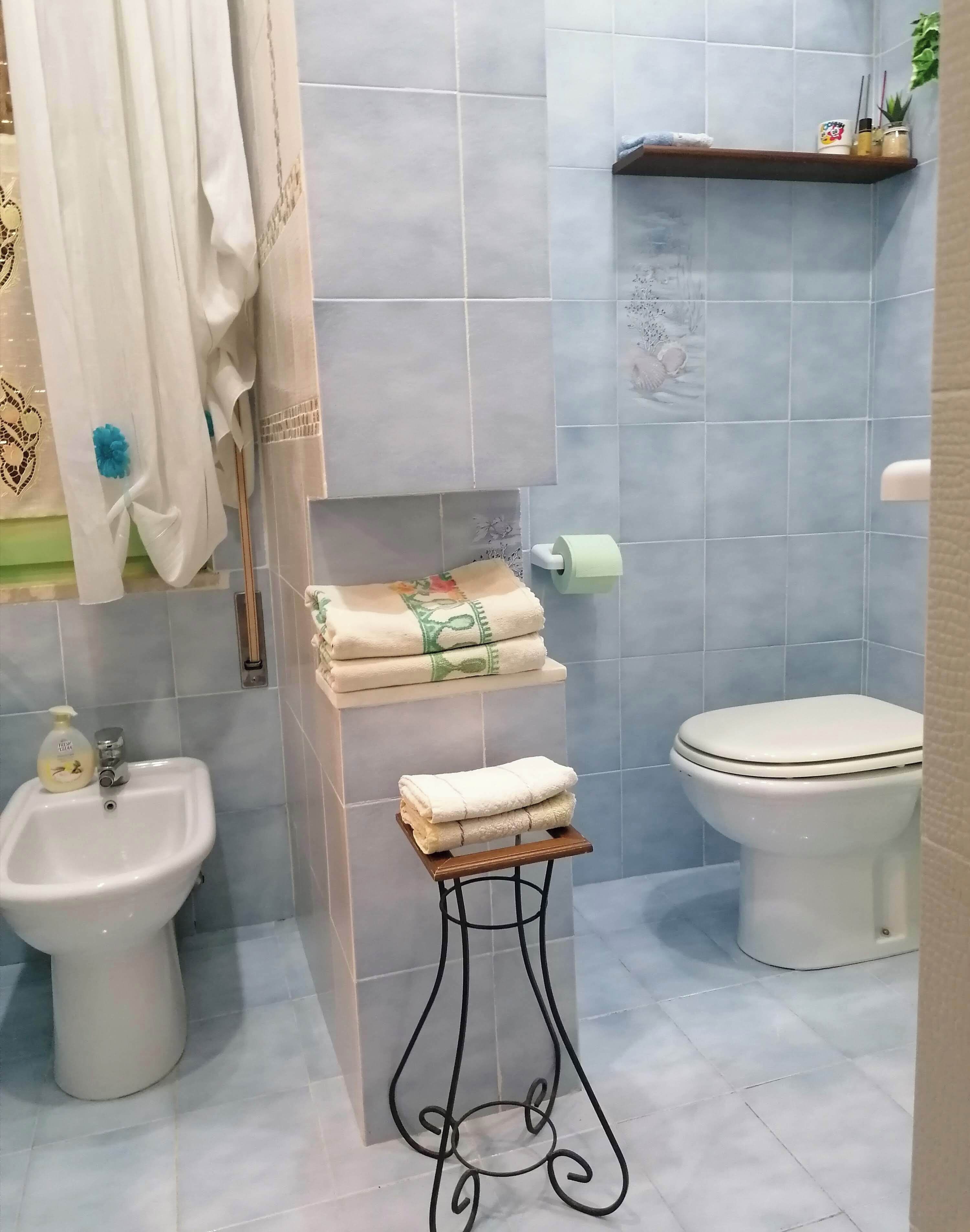 A bathroom with a bidet, toilet, and sink. There are towels stacked on a shelf and a toilet paper holder on the wall.