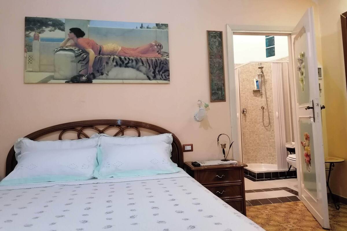 Une chambre à coucher avec un lit, une table de chevet, un tableau, une salle de bain avec une douche et des toilettes.