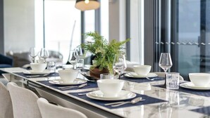 Mesa de comedor dispuesta con platos, cuencos y cubiertos blancos, una planta en maceta y una lámpara colgante moderna.
