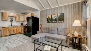 Ein modernes Wohnzimmer mit einem grauen Ecksofa, einem Glastisch und einer Küchenzeile mit Holzschränken und einem schwarzen Kühlschrank.