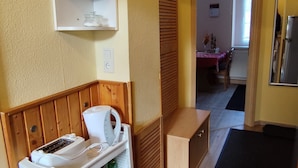 A compact kitchen with a refrigerator, a microwave, and a toaster. There is a wooden cabinet with a shelf above it holding jars. A wooden slatted partition is visible in the background.