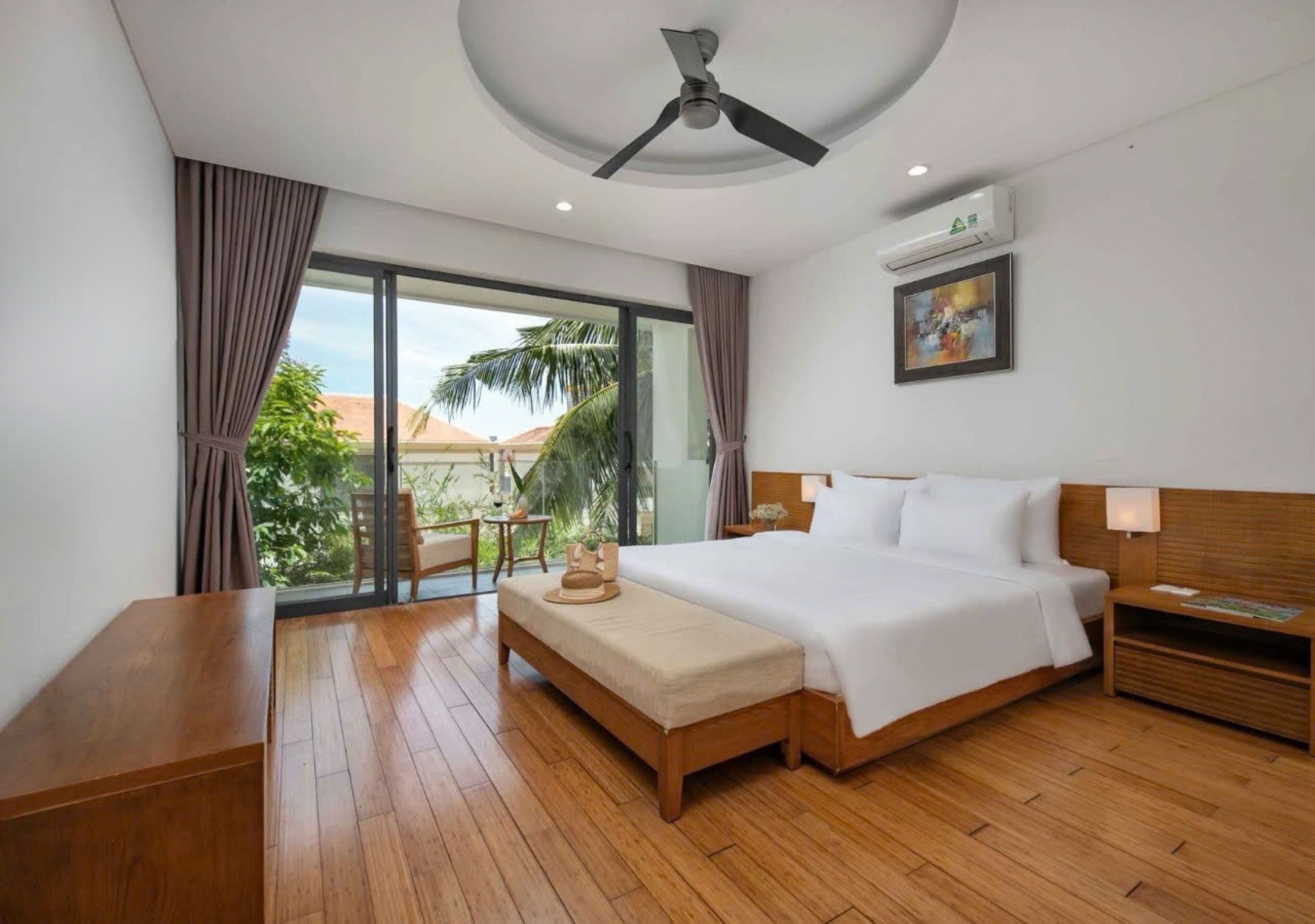 A hotel room with a large bed, a wooden bench, a ceiling fan, and a view of palm trees.