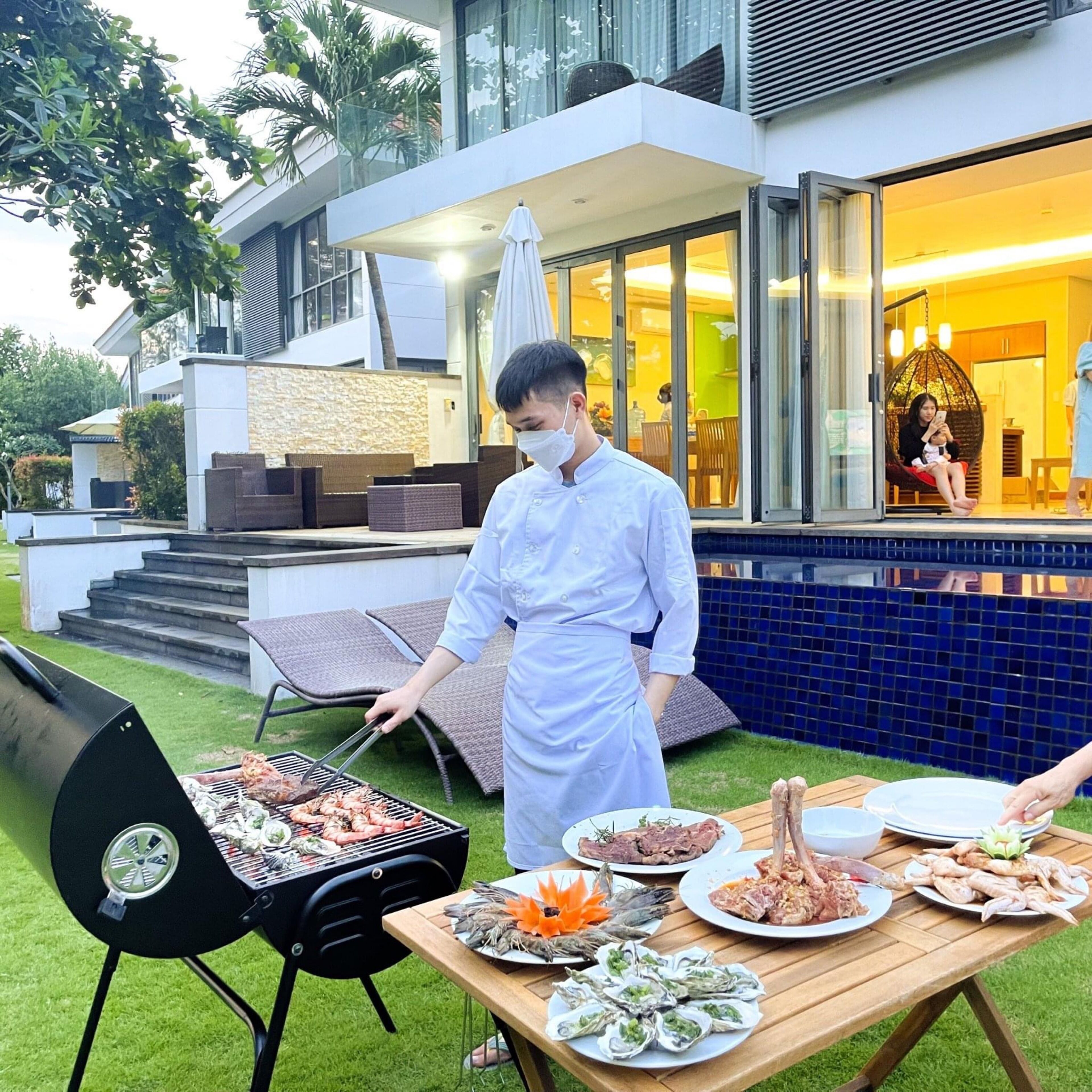 A chef grilling seafood outdoors in front of a modern house with a pool and a dining area visible inside.