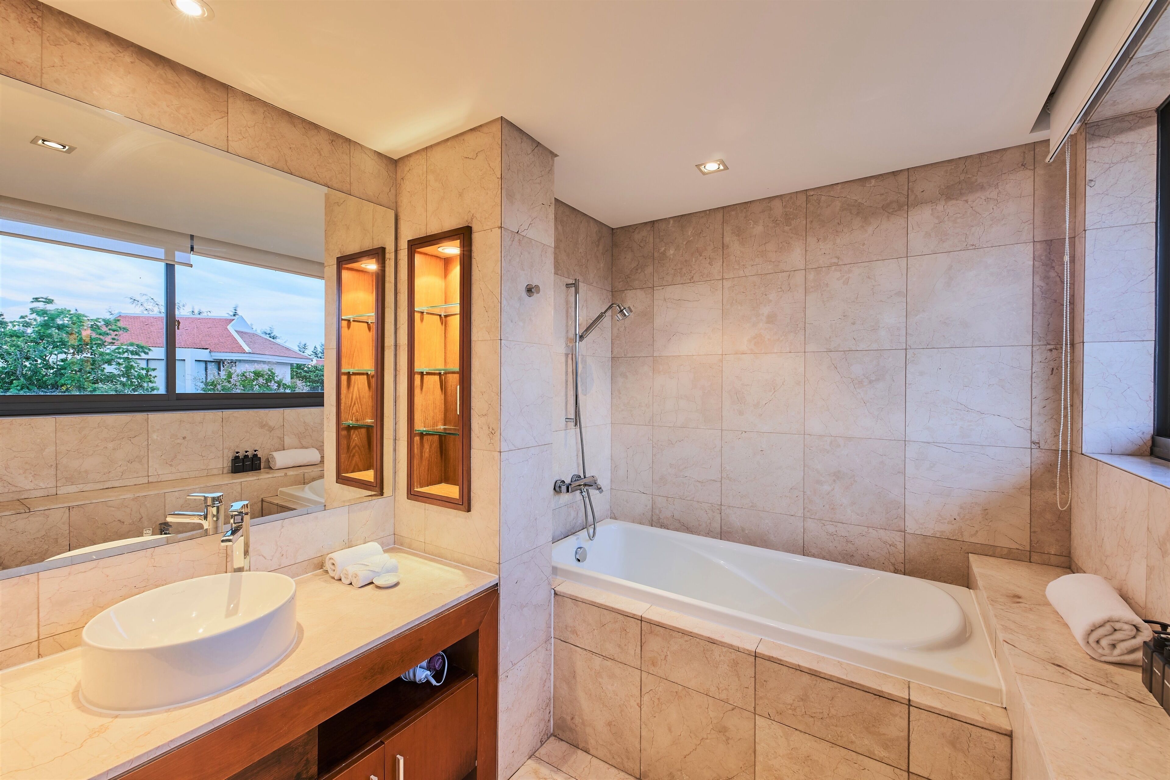 A modern bathroom with a large mirror, a sink, a bathtub, and a window offering a view of a building with a red roof.