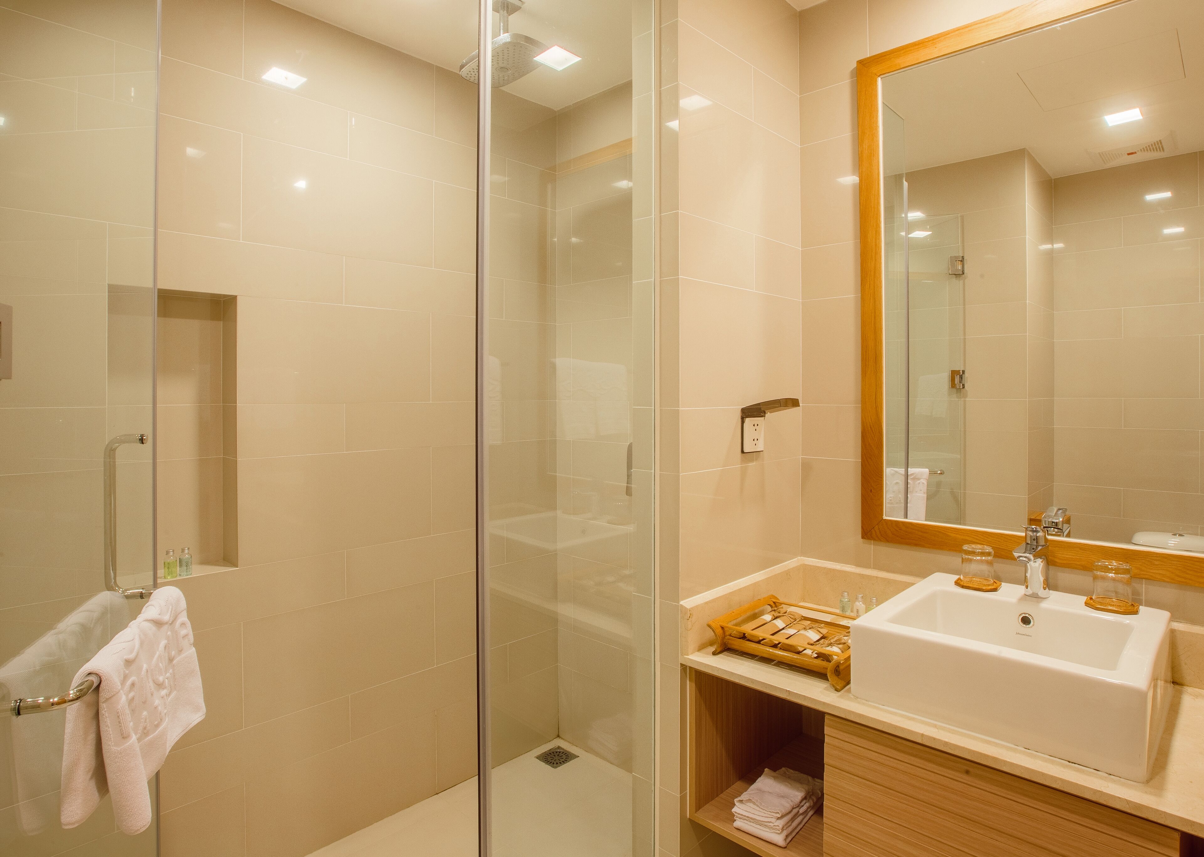 Un baño moderno con ducha envuelta en vidrio, un espejo grande y un lavamanos blanco sobre un mueble de madera.