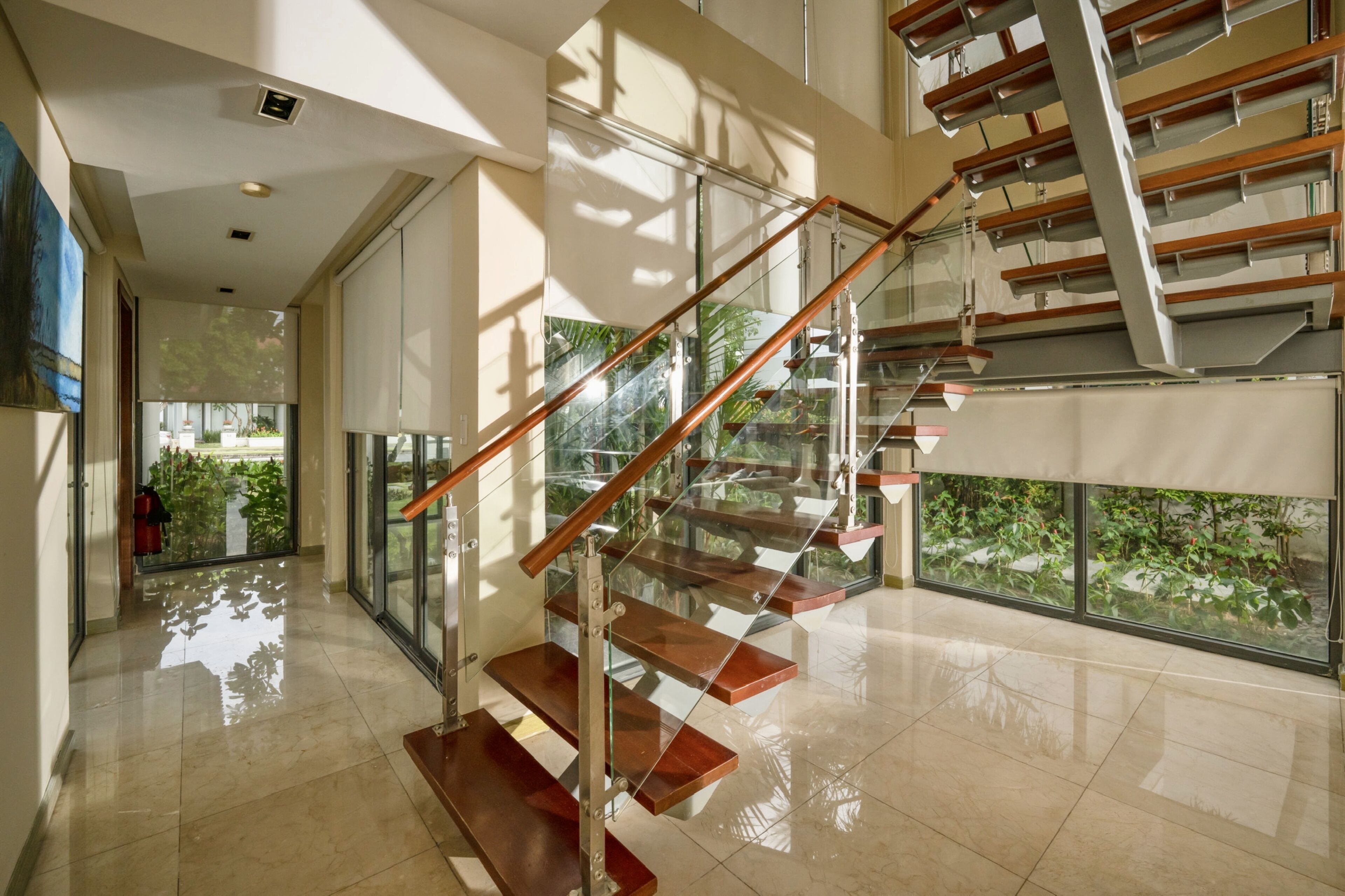 A modern interior with a glass staircase, polished floors, and large windows offering views of greenery.
