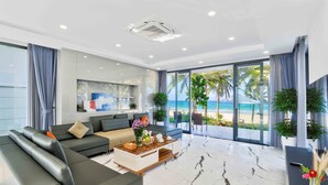 A modern living room with a large sectional sofa, a coffee table with decorative items, and a view of the beach through large glass doors. - The Ocean Suites (Da Nang)