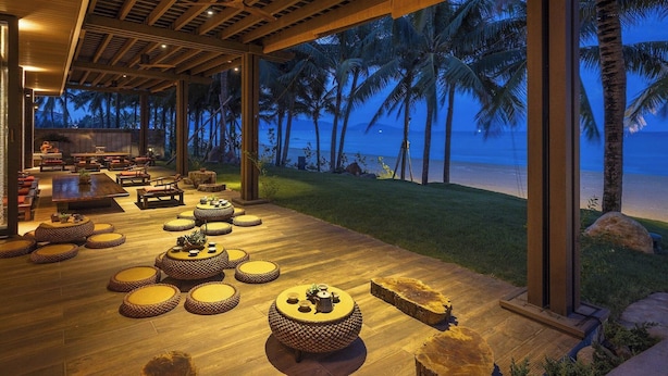 A spacious outdoor lounge area with wooden flooring, round seating, and large windows overlooking a beach and palm trees. - The Ocean Suites (Da Nang)
