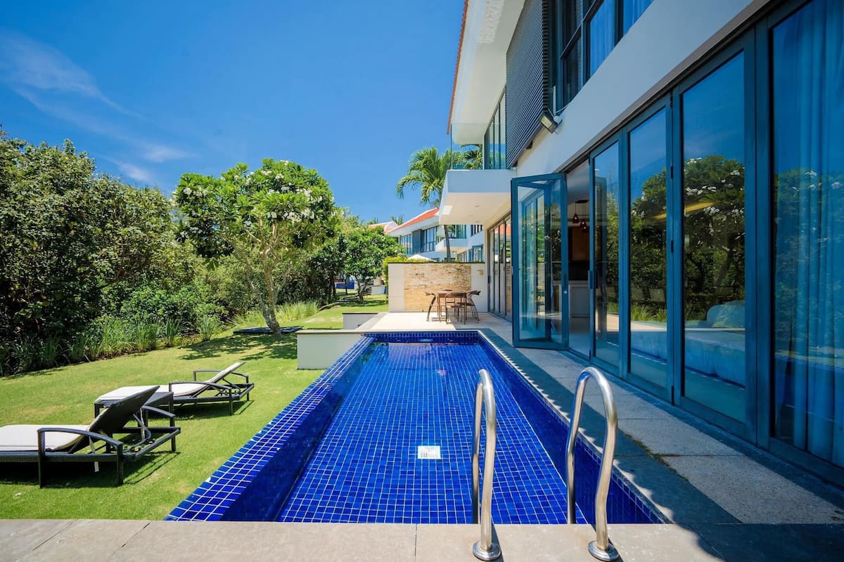 A modern outdoor swimming pool with a blue tiled floor, surrounded by lounge chairs and a glass-enclosed building.