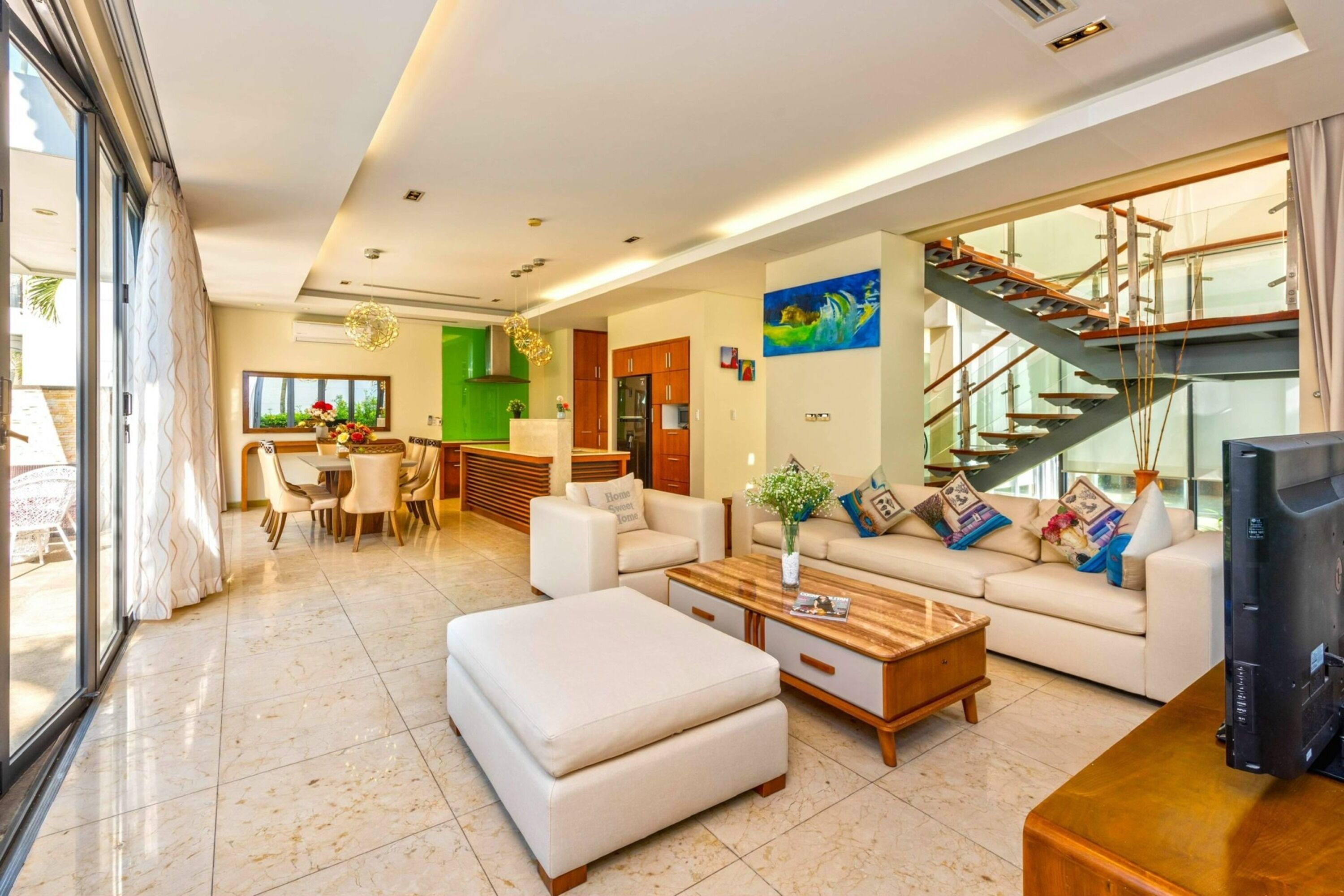 A spacious living room with a sofa set, a coffee table, and a television. There is a staircase with a glass railing leading to an upper level.